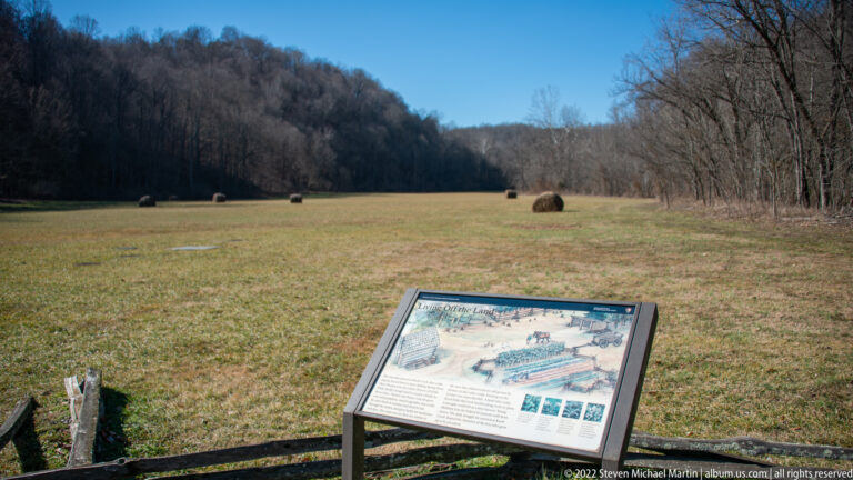 Round hay bales dot a valley field, with an angled interpretive panel in the foreground.