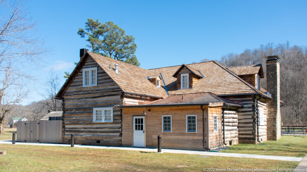 A reconstructed log cabin with a shingled roof, modern windows, and entry ramp stands on a grassy lawn.
