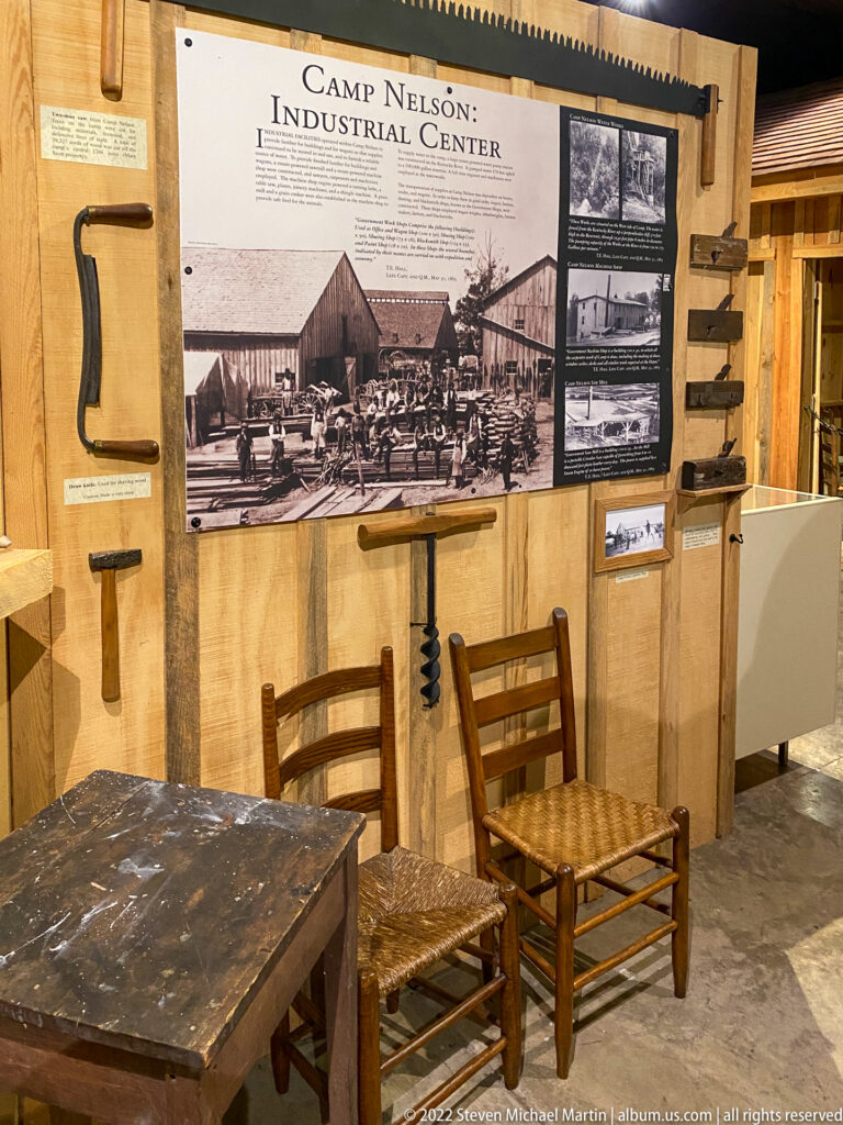 A Camp Nelson Industrial Center panel hangs above tools, two wooden chairs, and a worn worktable.