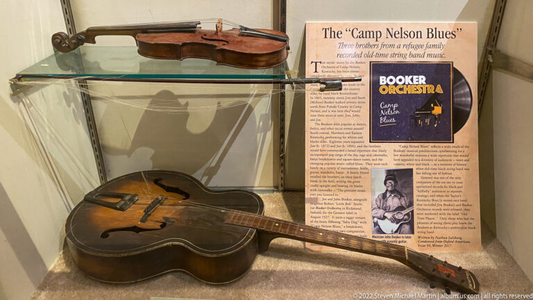 Two acoustic guitars sit on glass shelves beside a Camp Nelson Blues panel in a museum display case.