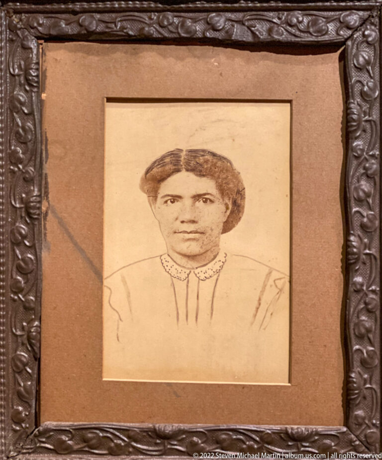 A forward-facing pencil portrait of a young person sits in a tan mat inside an ornate dark frame.