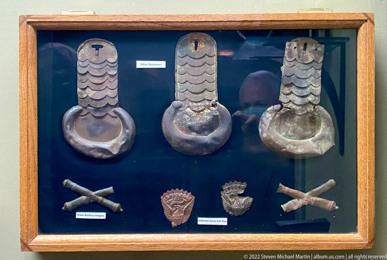 Three large metal fittings and several smaller objects are arranged in a labeled case against dark blue backing.