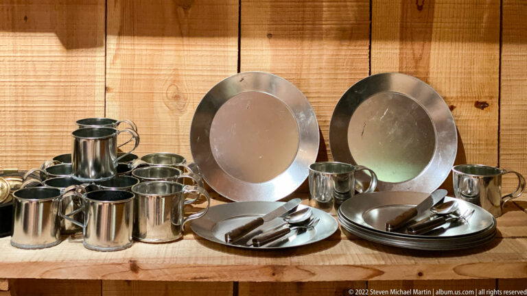 Metal cups, pitchers, plates, and utensils are arranged together on a wooden museum shelf.