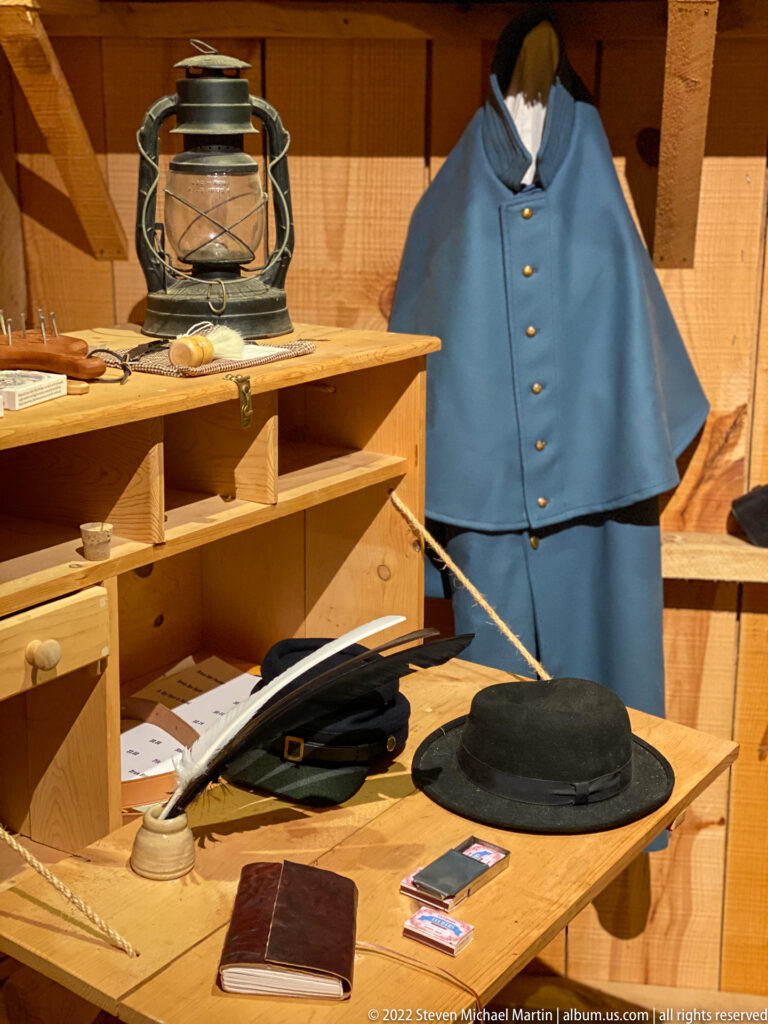 A blue buttoned garment hangs above a table with hats, papers, and small objects in a wooden display.
