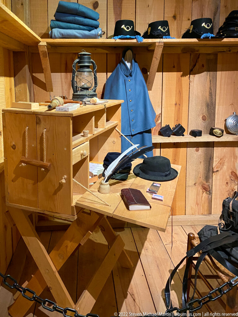 A wooden display holds a blue garment, stacked hats, folded textiles, a lantern, and other objects behind a chain barrier.