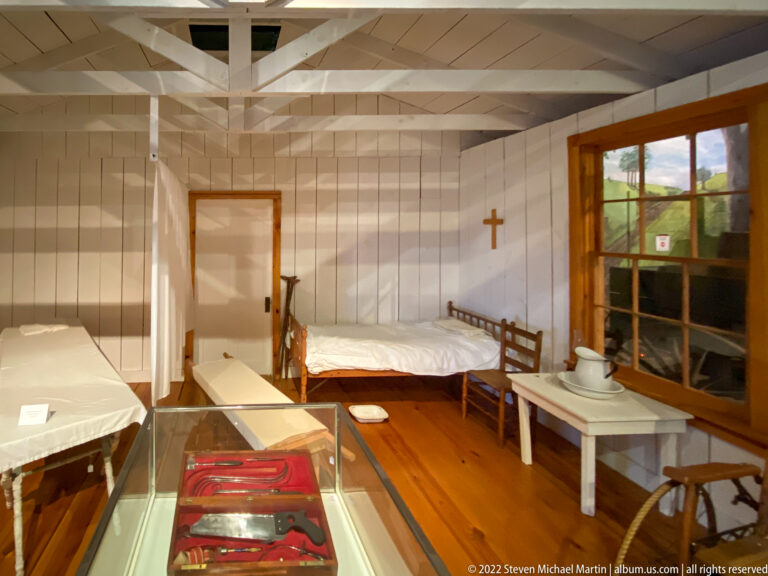Two narrow beds, a privacy screen, washstand, and wall cross furnish a reconstructed room with an exhibit case in front.