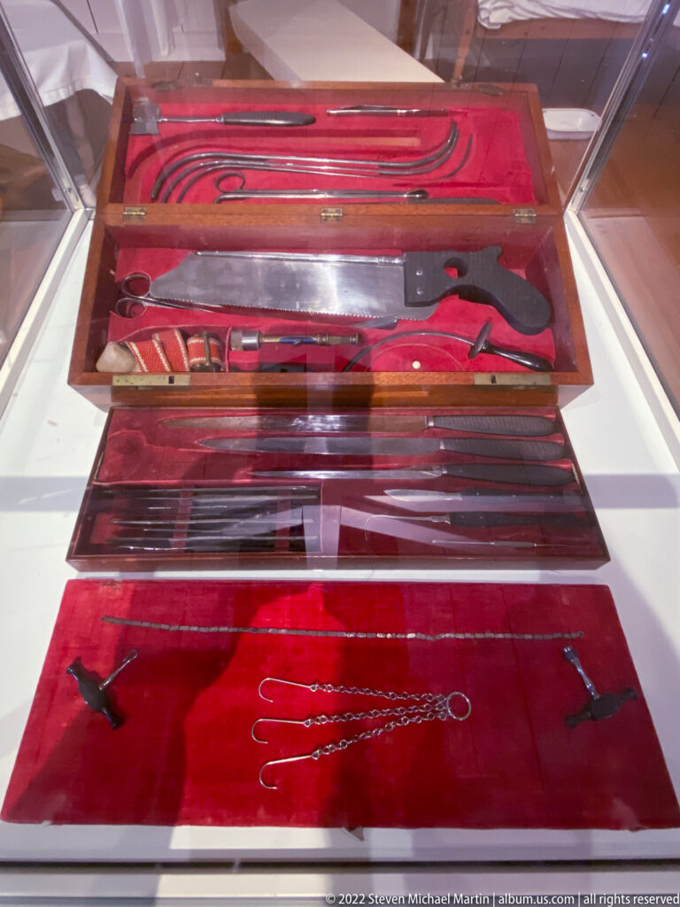 Long metal instruments with dark handles lie in red-lined fitted trays inside a glass display case.