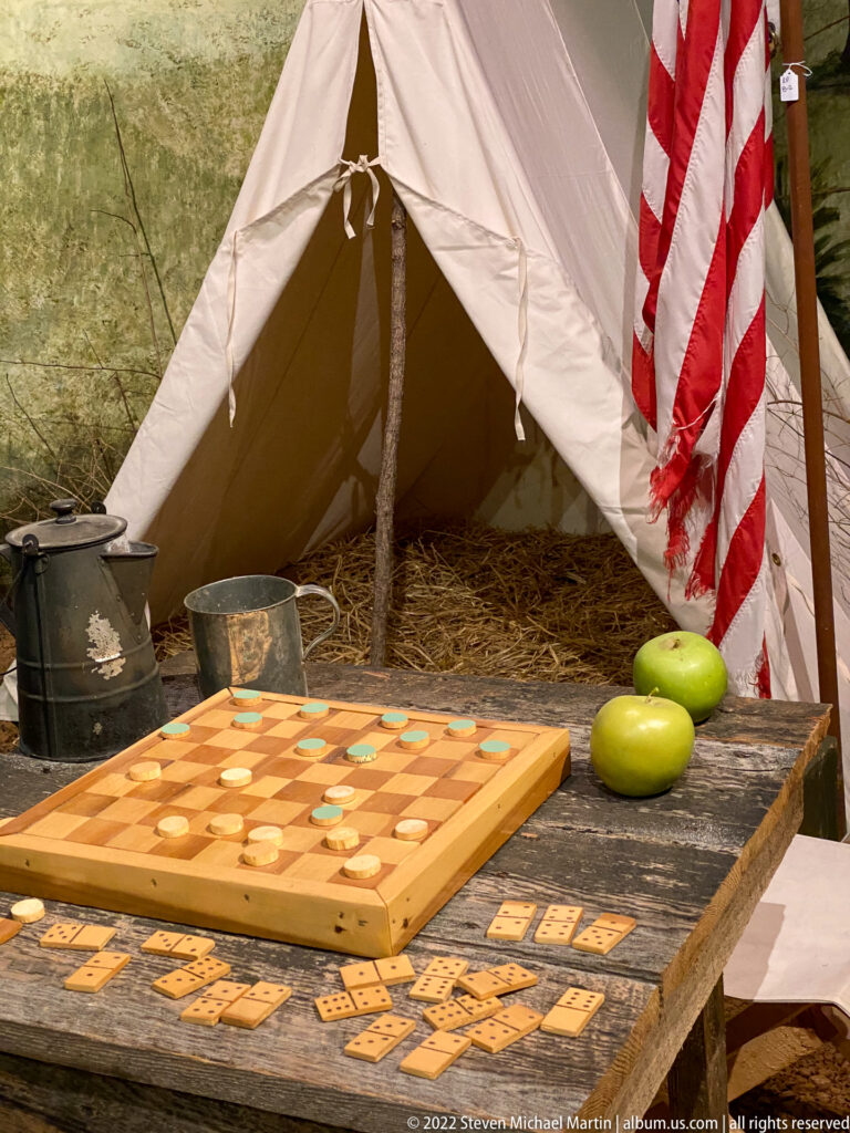 A checkerboard and scattered pieces share a rough table with tin pots beneath a U.S. flag beside a canvas tent.