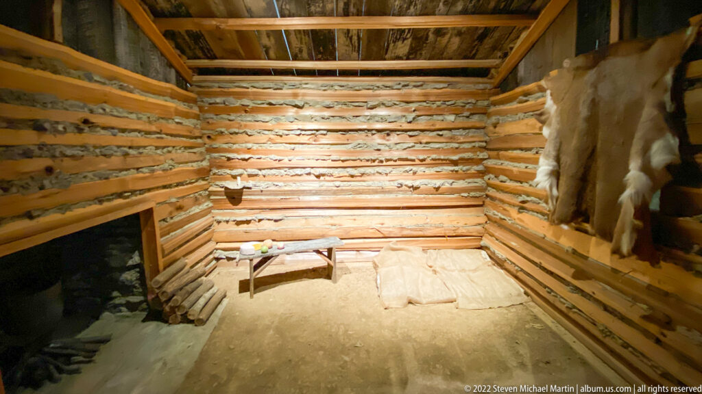 A small log room with chinked walls contains a low table and furnishings, with an animal hide hanging at right.