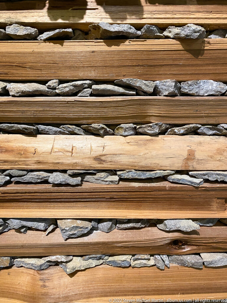 A close view shows horizontal timber courses separated by flat gray stones in a log wall.