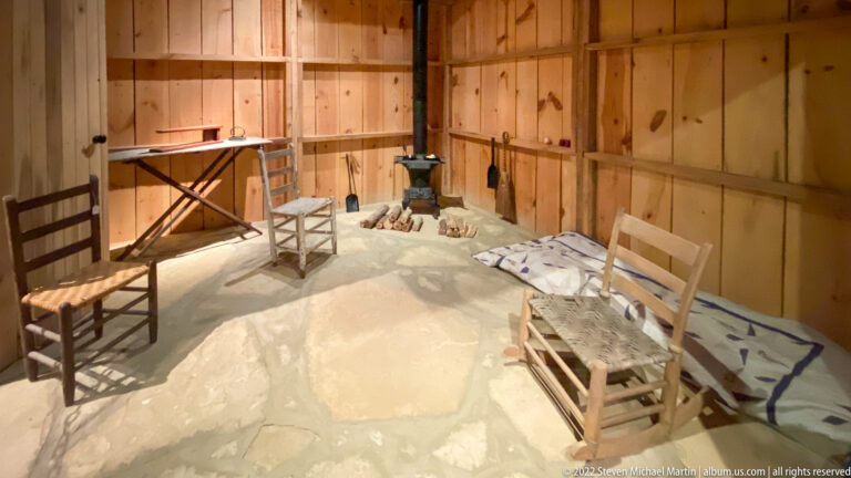 A small wooden room contains chairs, a narrow table, a bed, and a black iron stove.
