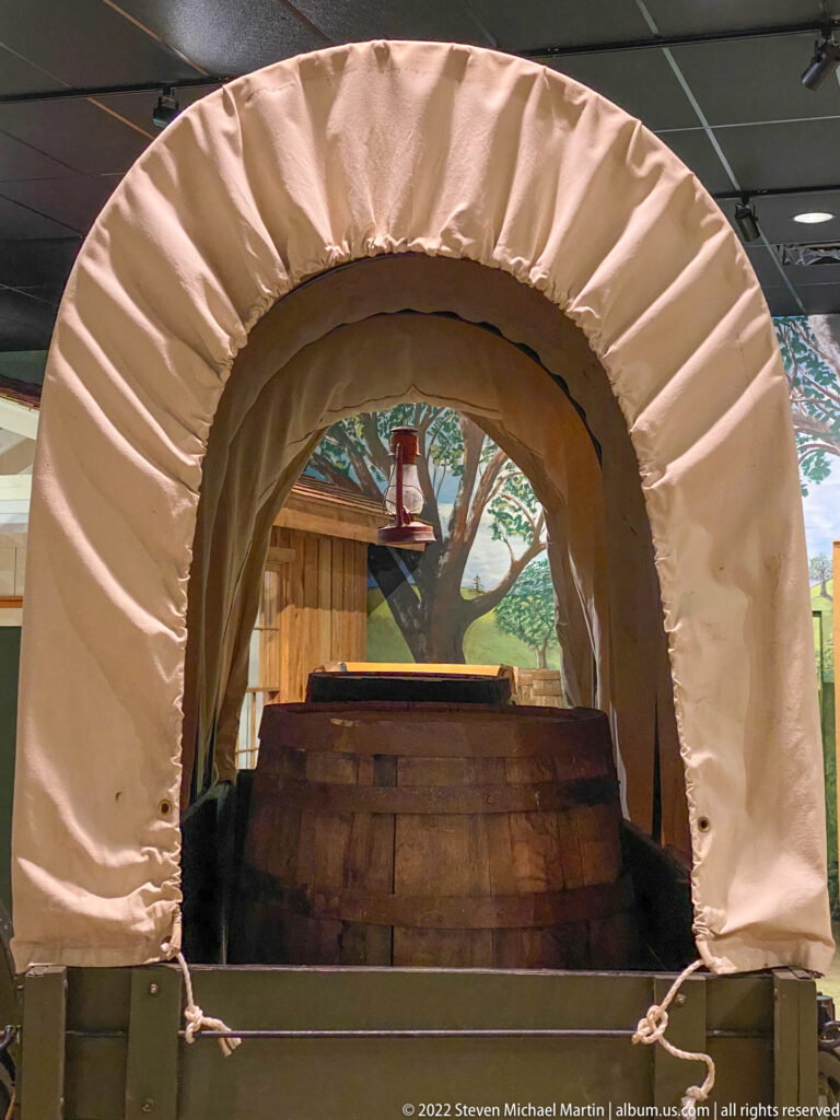 A pale canvas hood arches over a wooden wagon bed behind a rope barrier in a museum gallery.