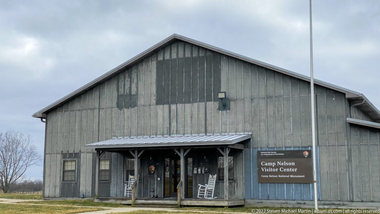 A large gray gabled building marked “Camp Nelson Visitor Center” stands beside a flagpole and winter lawn.