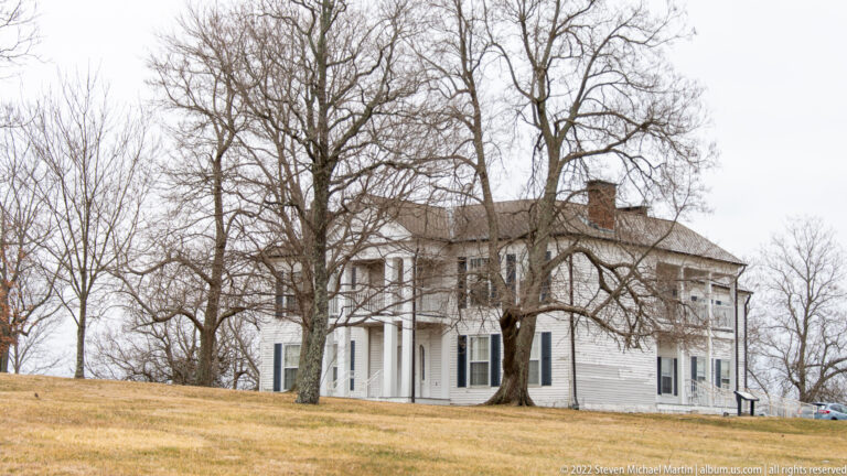 A white two-story house with a columned porch stands on a lawn behind leafless trees.