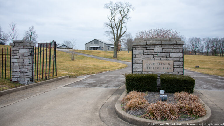 A paved entrance passes an open iron gate and a stone sign reading “Camp Nelson Heritage Park.”