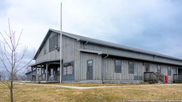 A long gray visitor-center building has a covered entrance, side windows, and a ramp across a winter lawn.