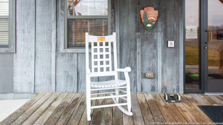 A white rocking chair sits on a weathered porch beneath an NPS emblem beside a glass entrance door.