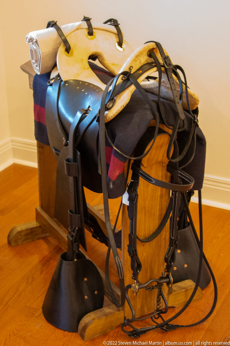 A dark leather saddle with straps and metal fittings rests on a wooden support beneath a patterned blanket.
