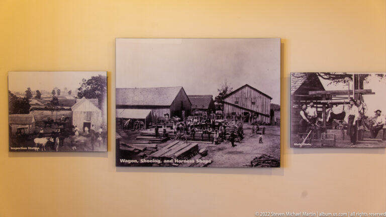 Three archival photographs hang on a wall; the central image is labeled “Wagon, Shoeing, and Harness Shops.”