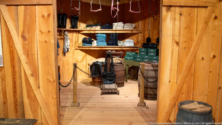 A wooden exhibit room contains folded clothes, hanging items, boxes, containers, and a small wheeled object behind a chain barrier.