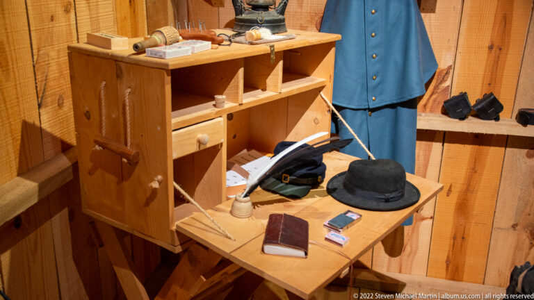 A blue coat hangs beside shelves and a wooden table with a black hat, books, papers, and small objects.