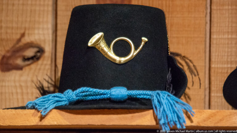 A black felt hat bears a brass bugle-shaped badge and a bright blue cord around its crown.