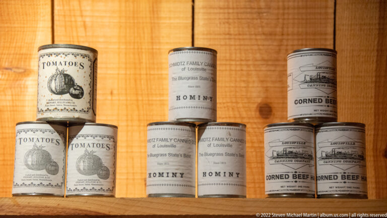 Cans labeled tomatoes, hominy, and corned beef are stacked on a wooden shelf.