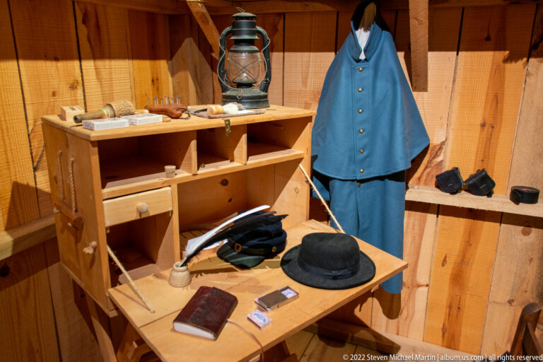 A wooden display room contains a blue coat, lantern, black hat, books, straps, and equipment arranged on shelves and a table.