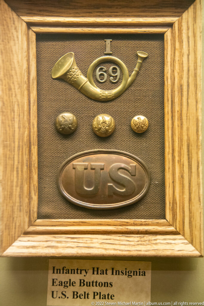 A framed display groups a brass “69” insignia, three eagle buttons, and a U.S. belt plate.