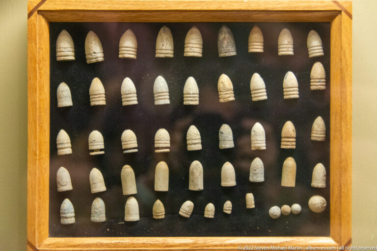 Rows of lead projectiles in varied sizes and shapes are displayed against a dark backing in a wooden case.