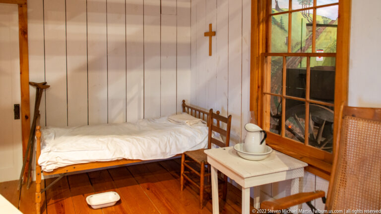 A narrow bed, table with pitcher and basin, chair, and wall cross furnish a small room beside a multi-pane window.