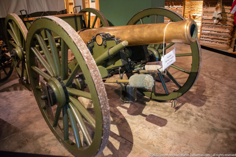 A field cannon on a green carriage stands indoors, with its spoked wheels and barrel prominent in the foreground.