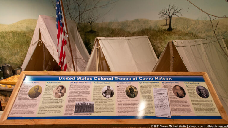 A United States Colored Troops interpretive panel stands before reconstructed canvas tents and an American flag.