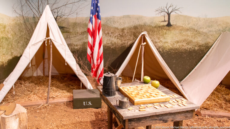 Canvas tents surround a table, cooking pots, and a box marked U.S., with an American flag between them.