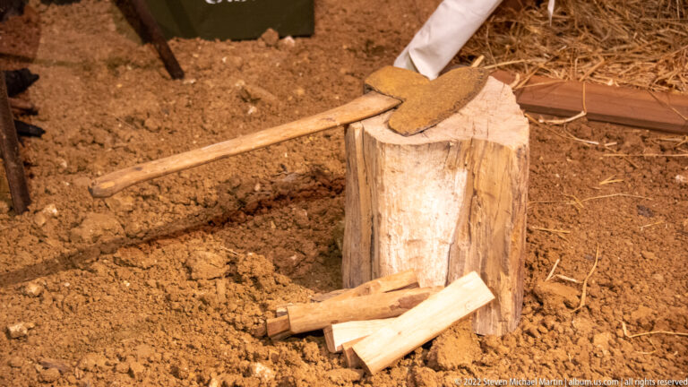 A long-handled axe lies across a tree stump beside split logs on straw-covered ground.