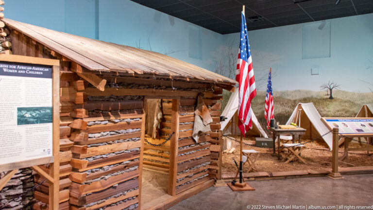A reconstructed log shelter stands beside interpretive panels, flags, tents, and a display table in a museum.