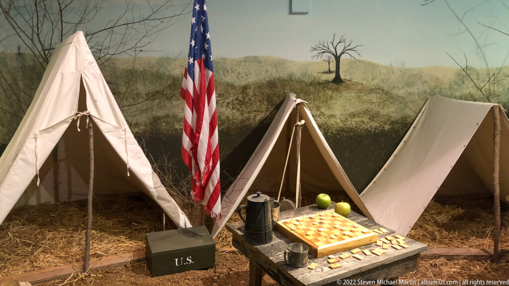 Canvas tents and a U.S. flag frame a table with a checkerboard and tin vessels in a museum scene.