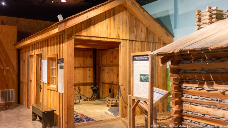 A small timber cabin with an open doorway stands beside a reconstructed log shelter and exhibit panels.