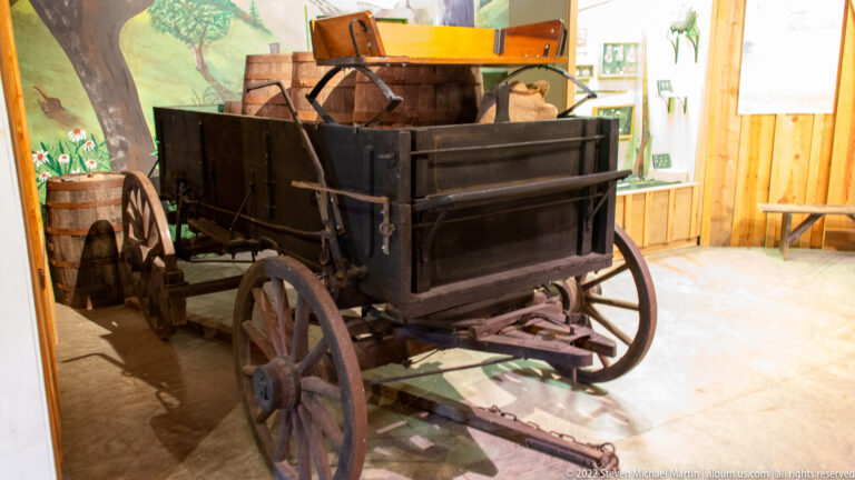 A dark wooden wagon with high sides, a raised seat, and spoked wheels stands in a museum exhibit.
