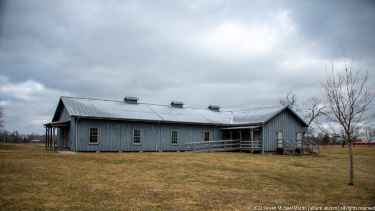 A long gray-blue building with a metal roof and rows of windows stands beyond a winter lawn.
