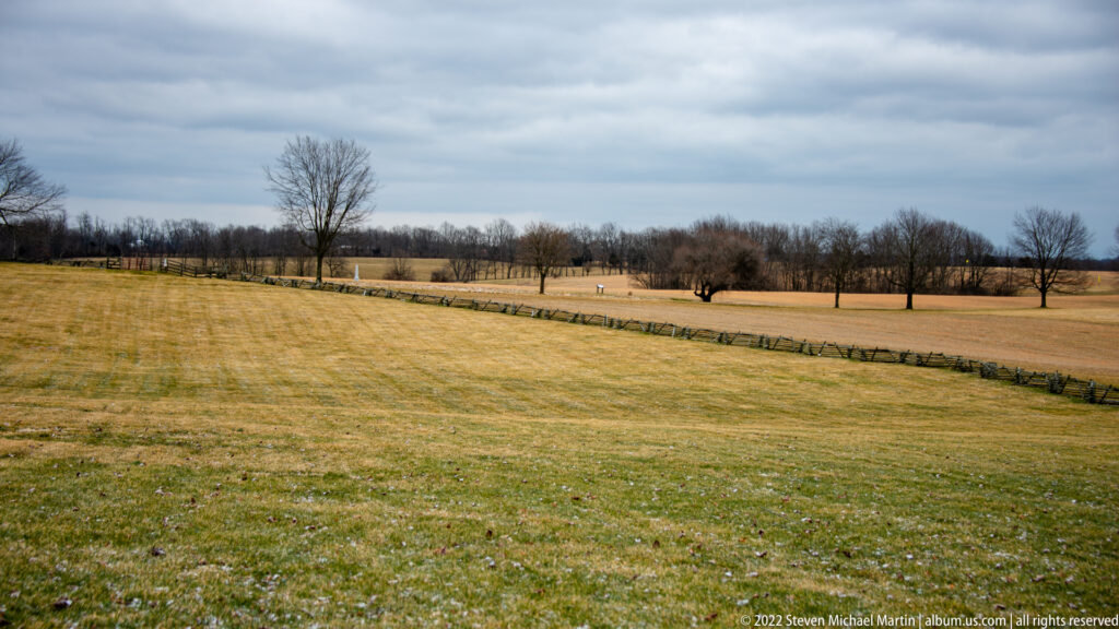 A low stone wall crosses a broad winter field toward scattered leafless trees and distant woods.