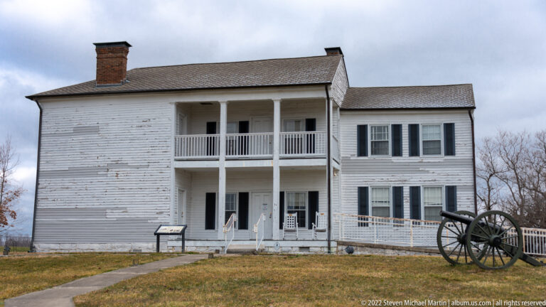A white two-story clapboard house has a double-height porch, dark shutters, and a cannon beside the lawn.