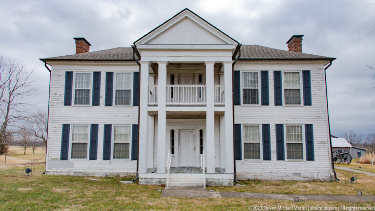 A white two-story house has a central portico, paired balconies, dark shutters, and brick chimneys.