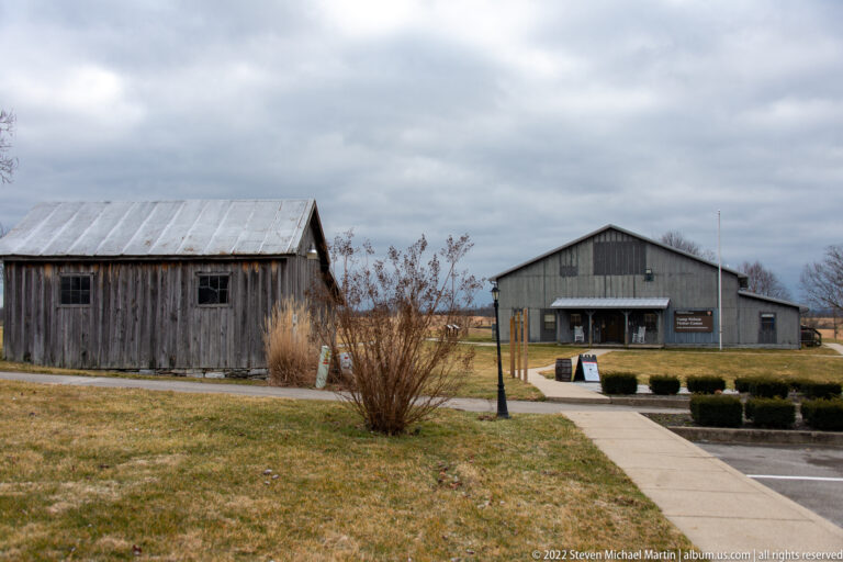 A small weathered outbuilding and a larger gabled building stand beside a paved path and winter lawn.