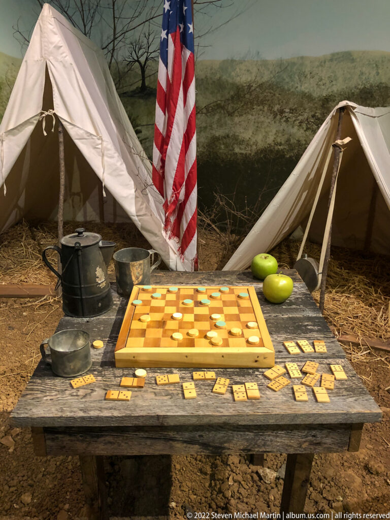 A checkerboard and scattered pieces sit among tin pots and cups on a wooden table beneath a U.S. flag.