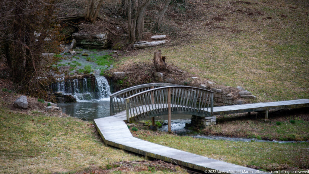 A small arched footbridge crosses a stream below a low waterfall, with a curving walkway in front.