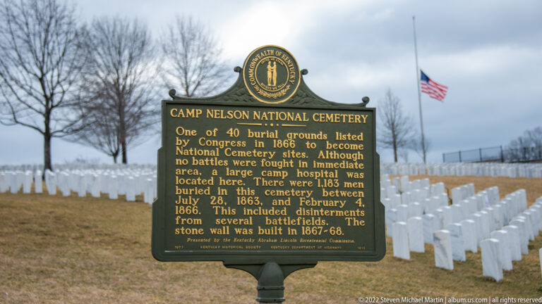 A Camp Nelson National Cemetery marker stands before rows of white headstones and a flagpole.