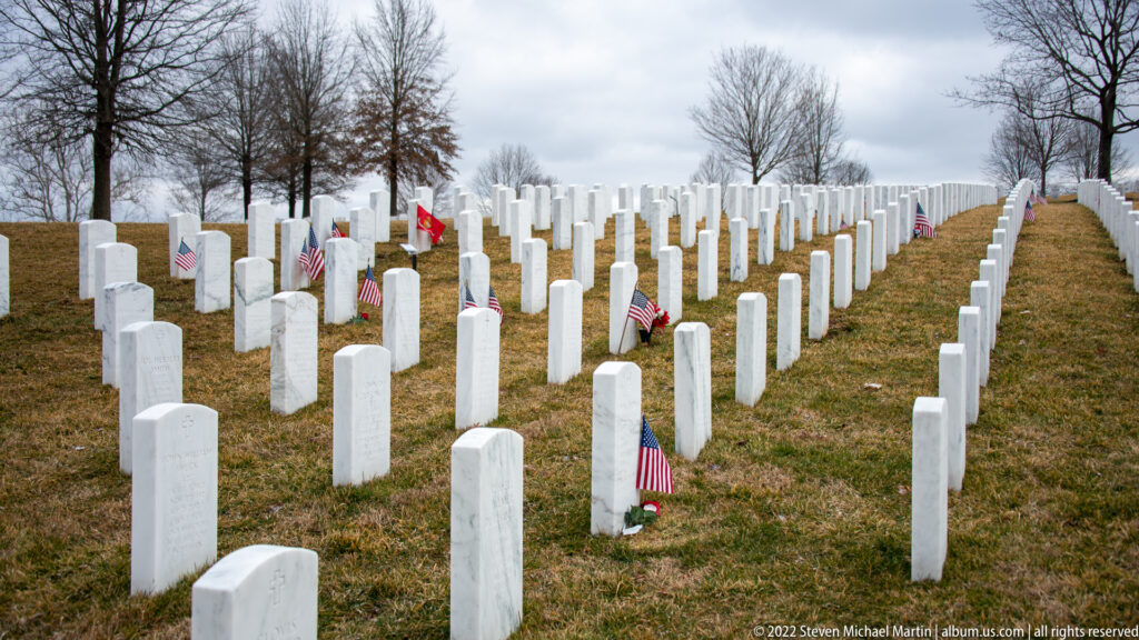 White headstones and small American flags line a gently sloping cemetery lawn beneath leafless trees.