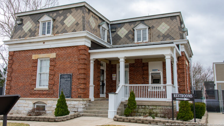 A red-brick building with a patterned roof and white-columned porch stands beside a restroom sign.