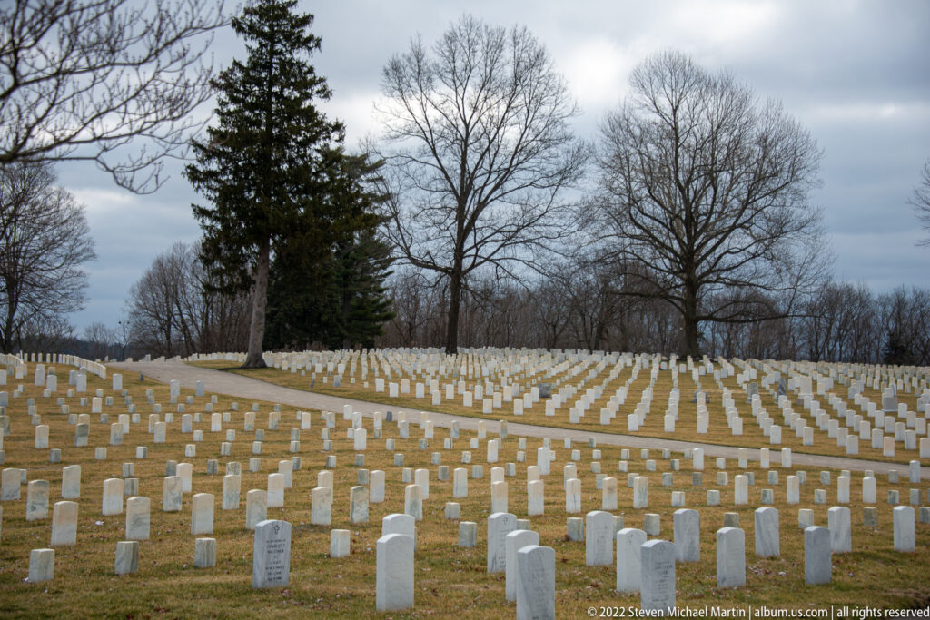 Rows of white headstones spread across a hillside beside a paved path and leafless trees.